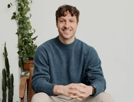 Homme souriant en pull bleu assis dans un cadre moderne, représentant une approche humaine pour la logistique IA.