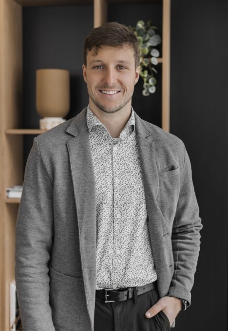Carl Beaudoin, expert en transport, souriant, vêtu d'une veste grise et d'une chemise à motifs, devant une étagère moderne.